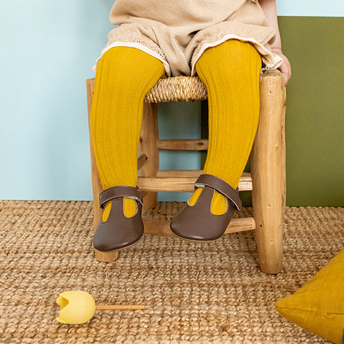 a small child sat on a wooden chair, shown from the waist down. The child is wearing yellow tights and brown t bar shoes. 
