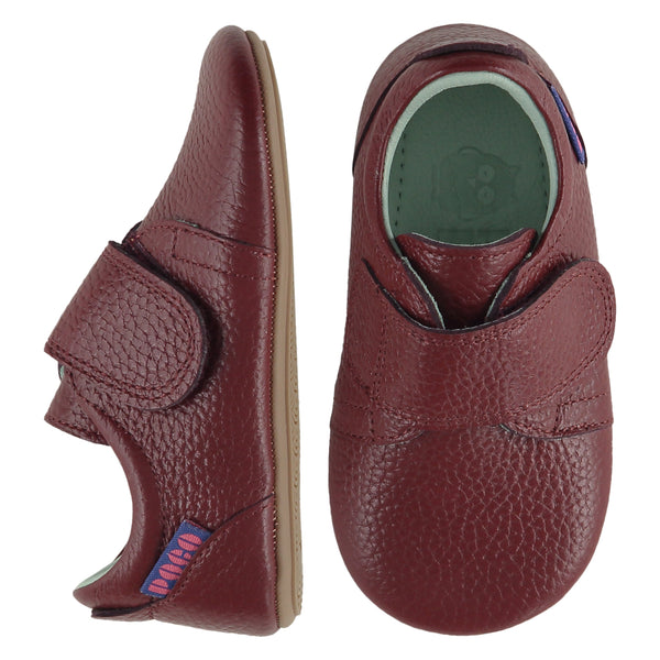 A pair of childrens adjustable strap shoes, one viewed from the top, one is a view from the side. They are in wine red leather with a large adjustable strap over the top of the foot. They have a  pale blue leather lining and insole. The sole is a natural gum colour.