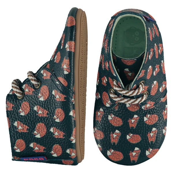 A pair of childrens lace up desert boots, one viewed from the top, one is a view of the side. They are in dark teal colour leather with striped laces and an all over print of foxes. They have a  pale blue leather lining and insole. The sole is a natural gum colour.