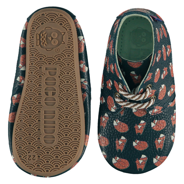 A pair of childrens lace up desert boots, one viewed from the top, one is a view of the sole. They are in dark teal colour leather with striped laces and an all over print of foxes. They have a  pale blue leather lining and insole. The sole is a natural gum colour.